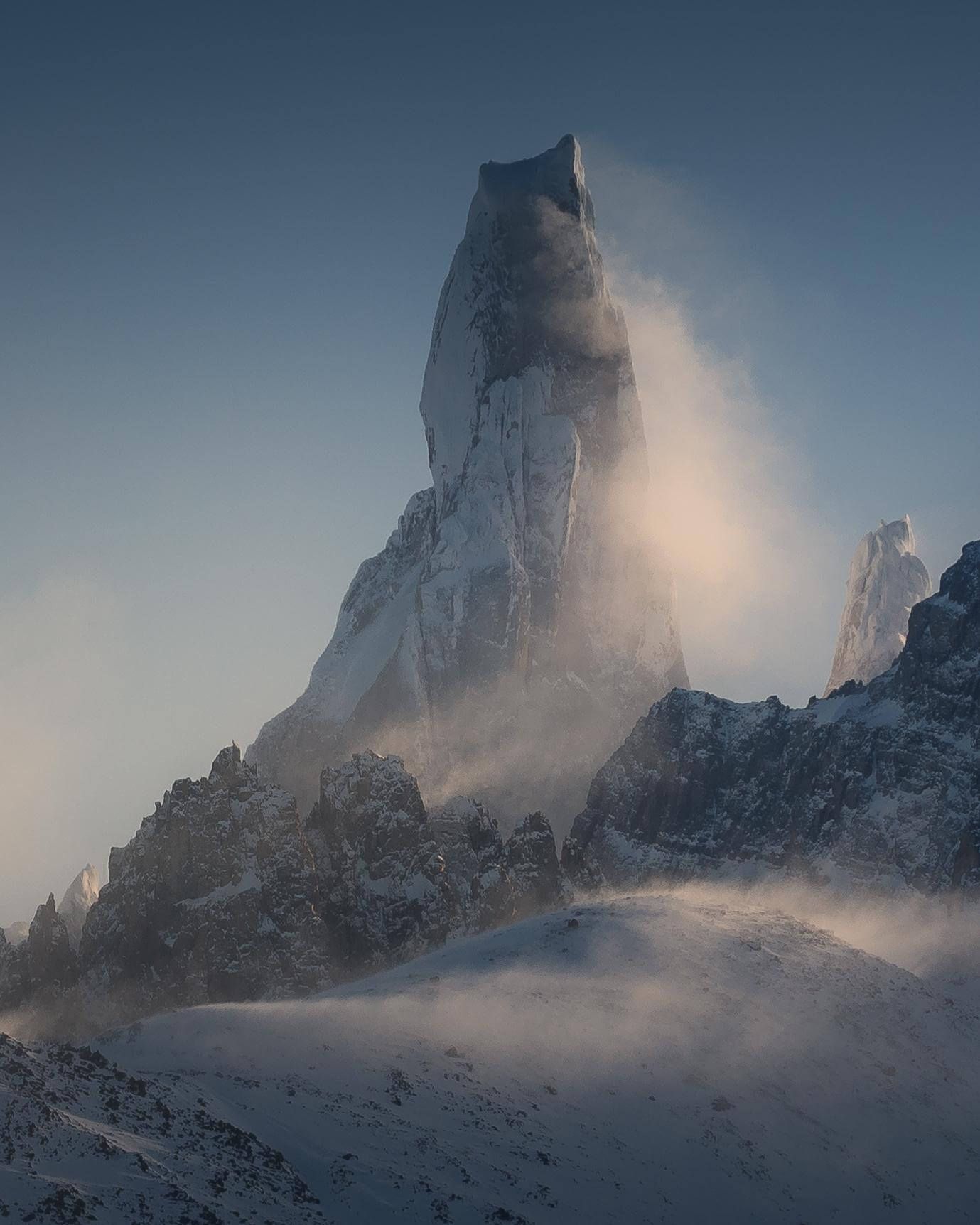 A lone jagged rocky mountain peak surrounded by swirling snow and mist against a clear blue sky.