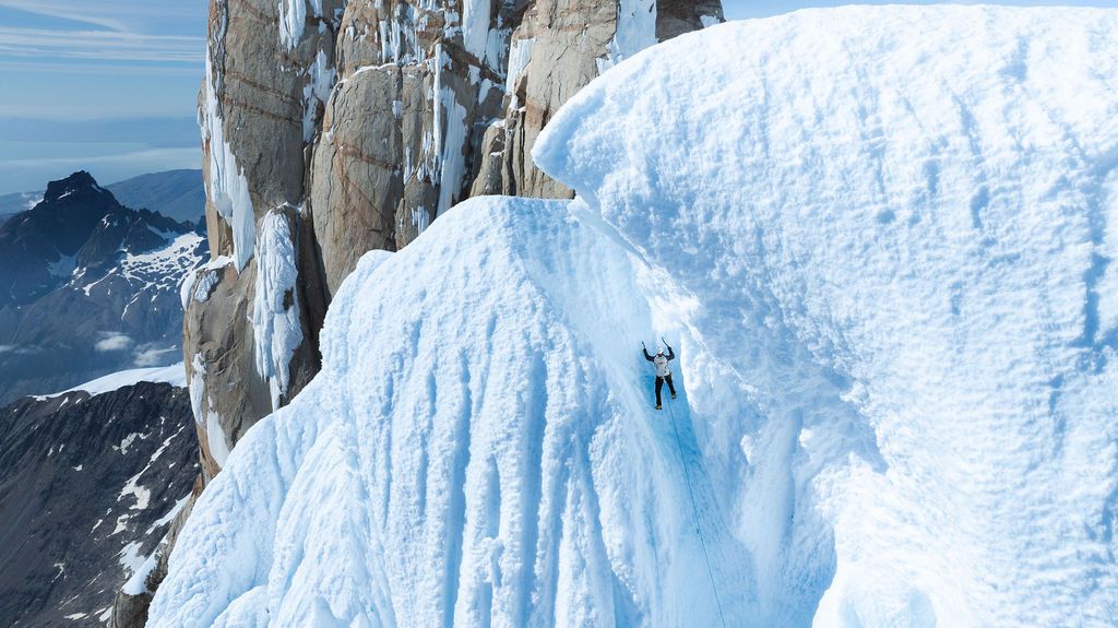 Climber ascending a steep ice-covered granite tower with dramatic mountain scenery in the background.