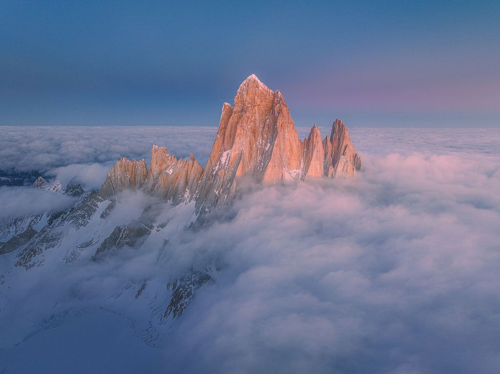 Jagged snow-capped mountain peaks rise dramatically above a sea of clouds at pink sunset in Patagonia.