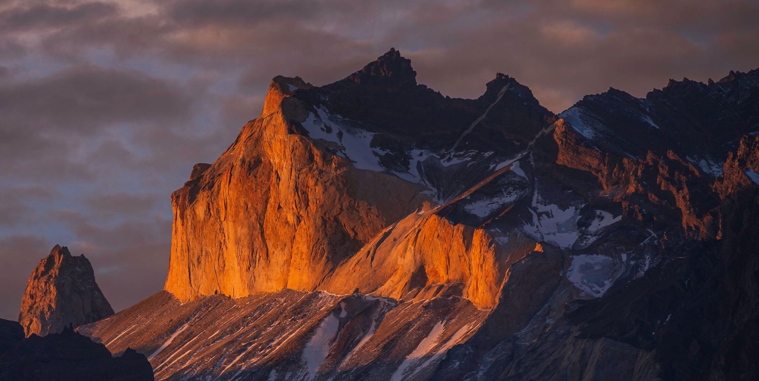 Golden alpenglow illuminates dramatic rocky mountain peaks at sunset with snow-capped summits against a stormy sky.