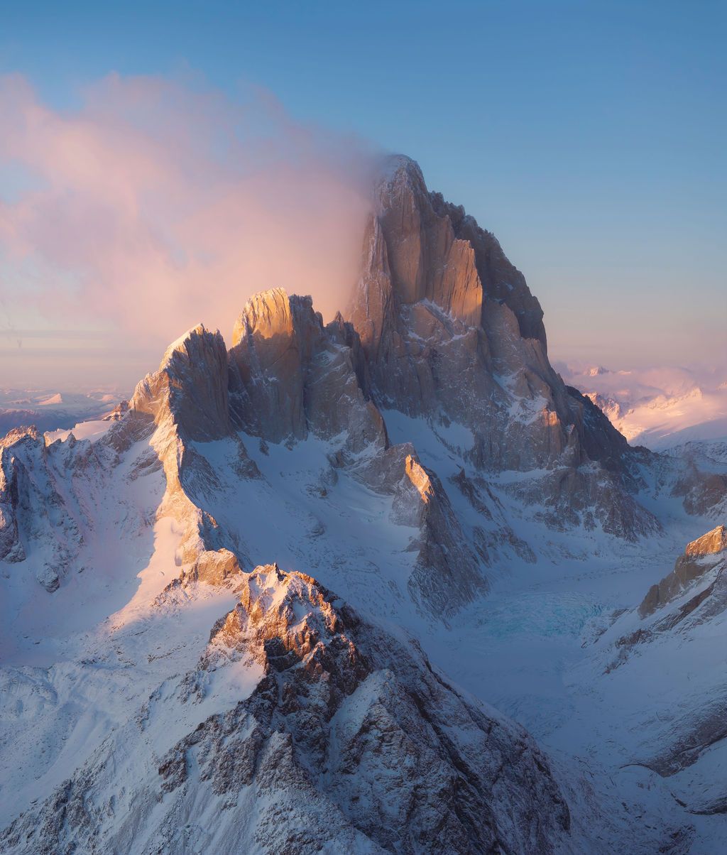 Aerial view of snow-covered mountain massif bathed in warm alpenglow light with dramatic pink clouds at sunset.
