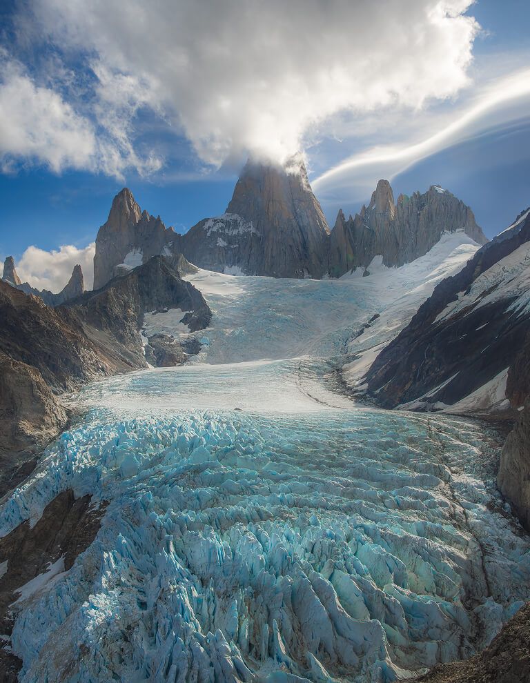 Aerial view of a massive blue glacier flowing between rocky peaks under dramatic clouds in Patagonia.