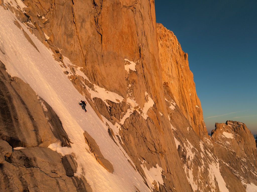 Climber ascends a steep snow slope beside a massive golden granite wall at sunrise in Patagonia.