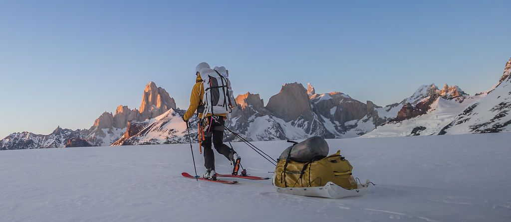 A skier pulling a sled across a snowy plain with rocky mountain peaks at sunset.