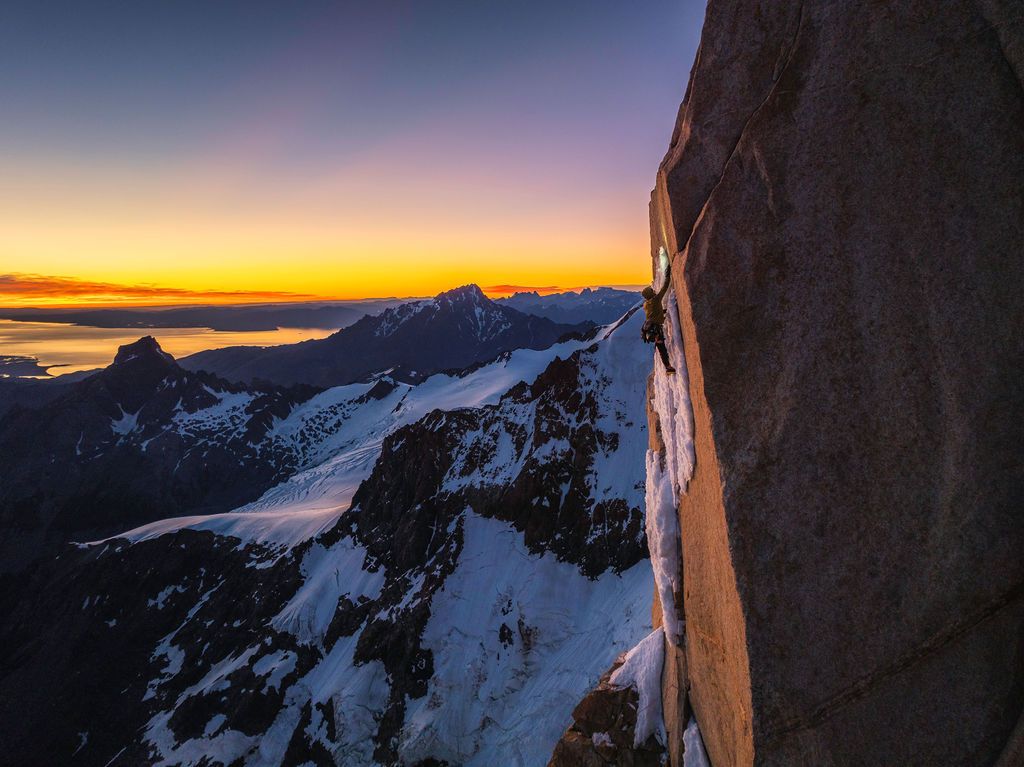Dramatic sunset view from a high mountain rock face with glowing orange sky over snow-capped peaks.