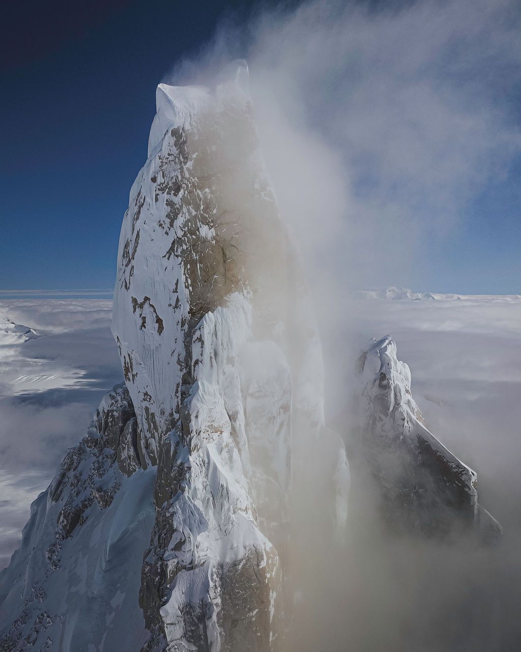 Snow-covered rocky mountain spire emerges through swirling clouds and wind against a clear blue sky.