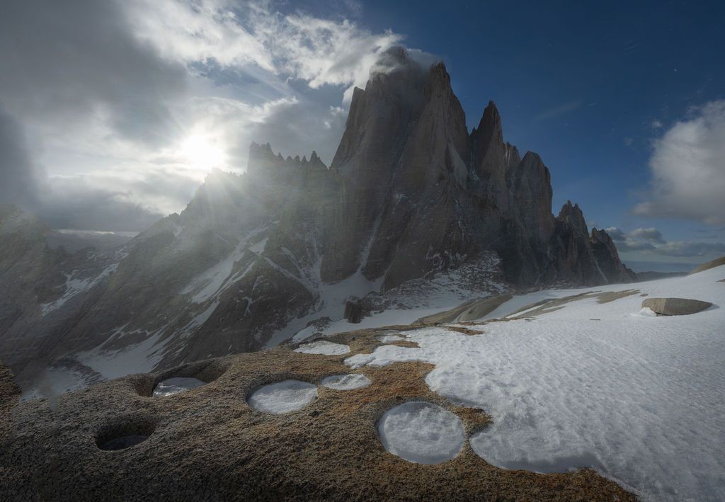 Sun rays burst over jagged rocky peaks with snow patches in the Dolomites, Italy.