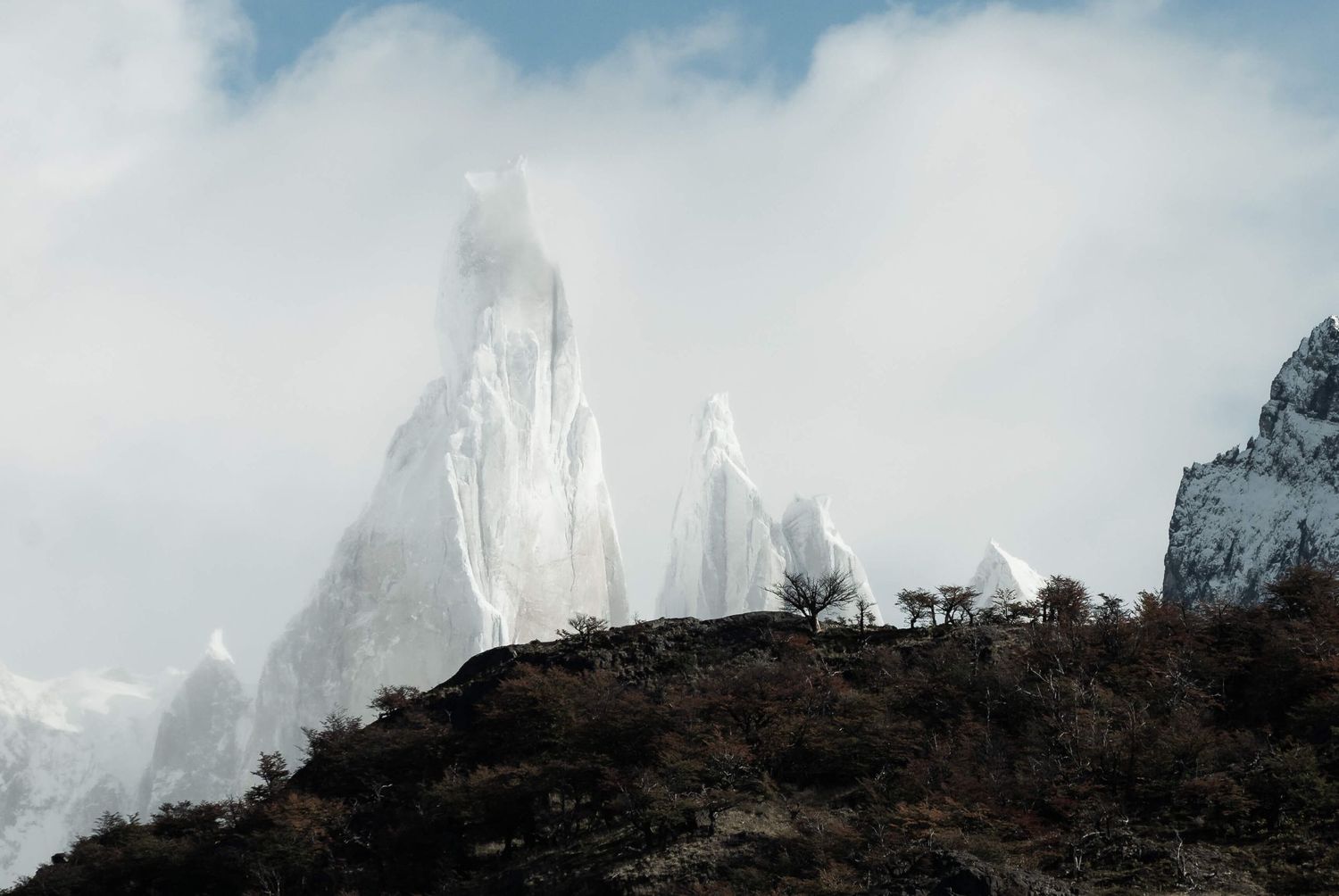 Snow-covered spire of Cerro Torre emerging through thick white clouds above a rocky hillside in Patagonia.