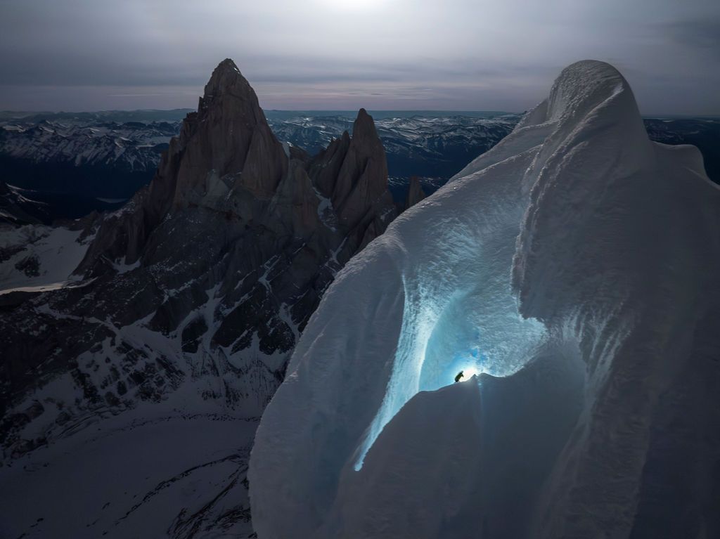 Climber with headlamp ascending a massive ice formation at dusk with jagged Patagonian peaks in the background.