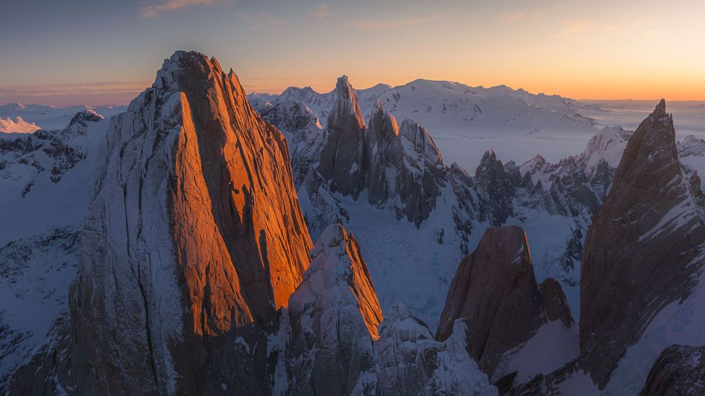 Dramatic aerial view of Cerro Torre and surrounding jagged peaks glowing orange at sunset in Patagonia.