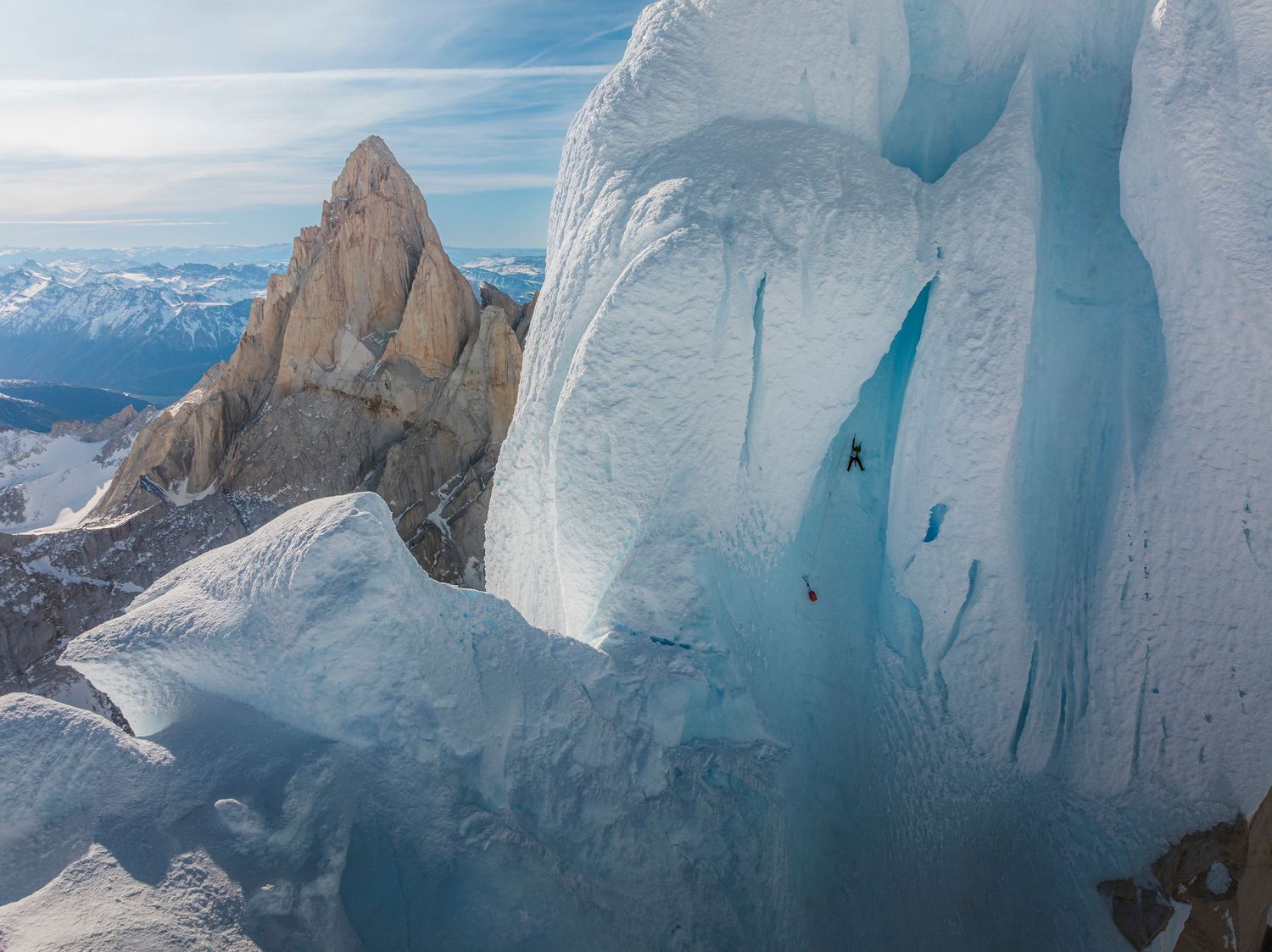 Two climbers scale a massive blue ice wall with a dramatic rocky mountain peak in the background under a clear sky.