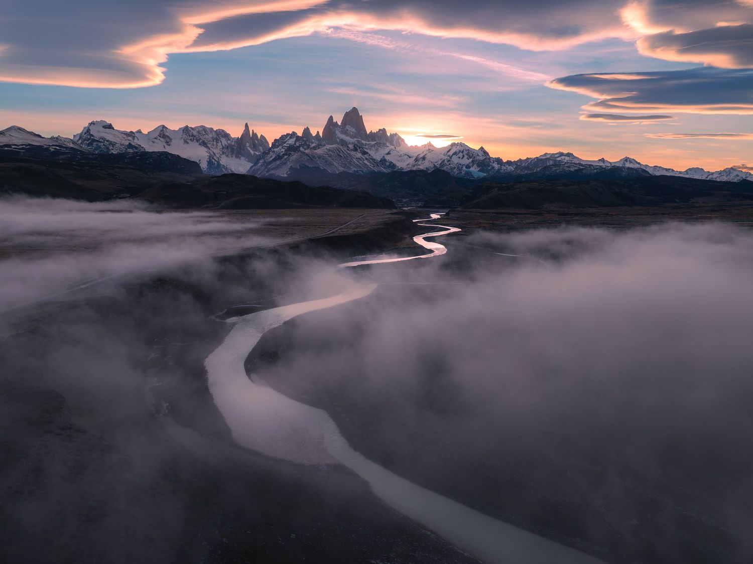 Aerial view of a winding river through misty Patagonian landscape with snow-capped Mount Fitz Roy at dramatic sunset.