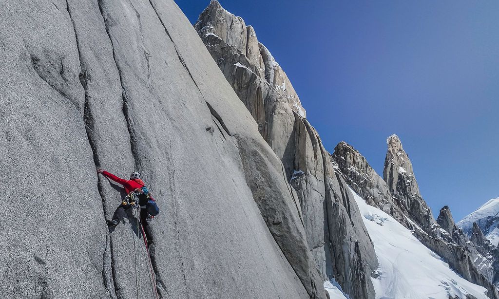 Rock climber in red jacket ascending a smooth granite slab with jagged snow-dusted Patagonian towers behind.