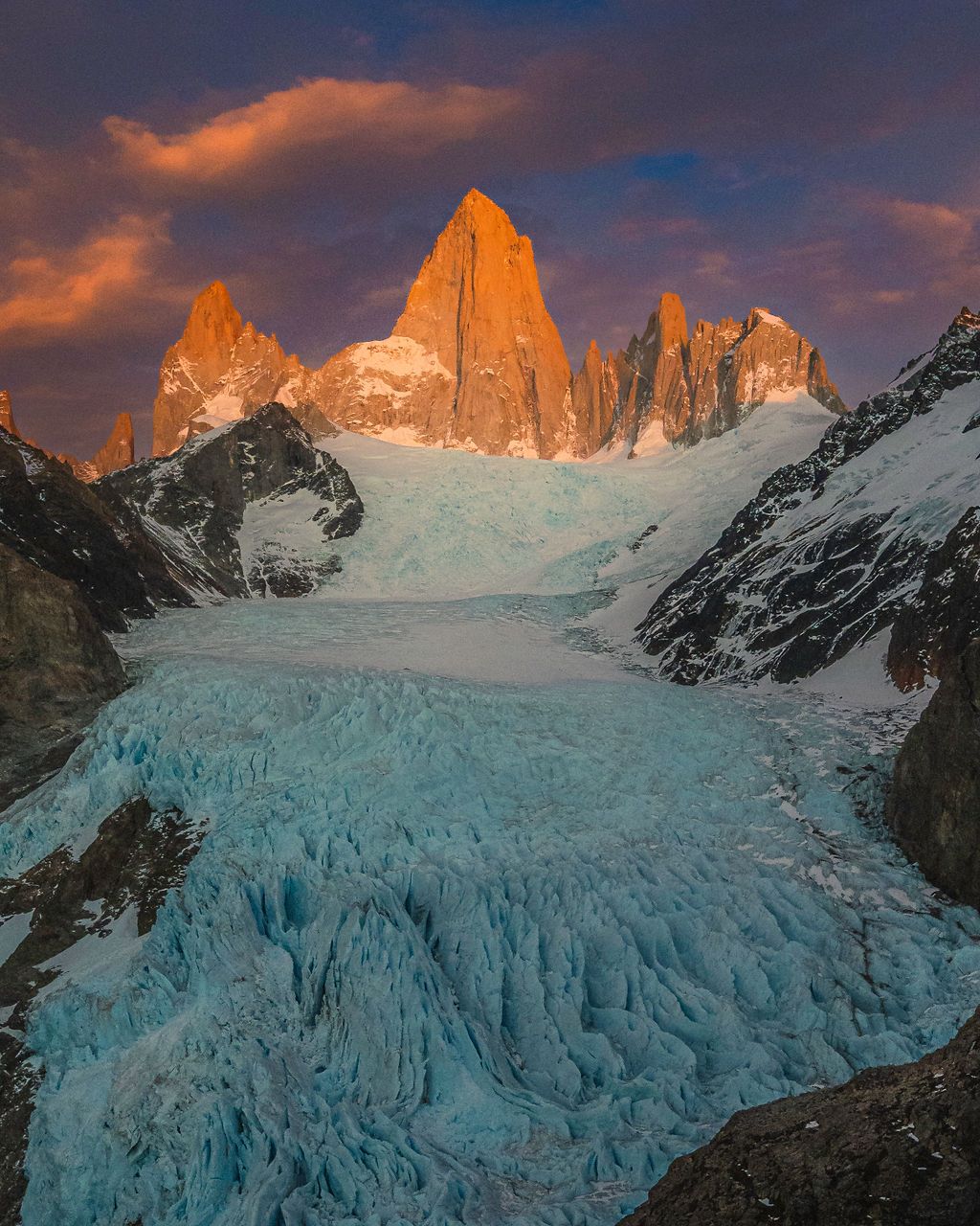 Glacial ice flows beneath the golden alpenglow on Fitz Roy's jagged peaks at sunrise in Patagonia.