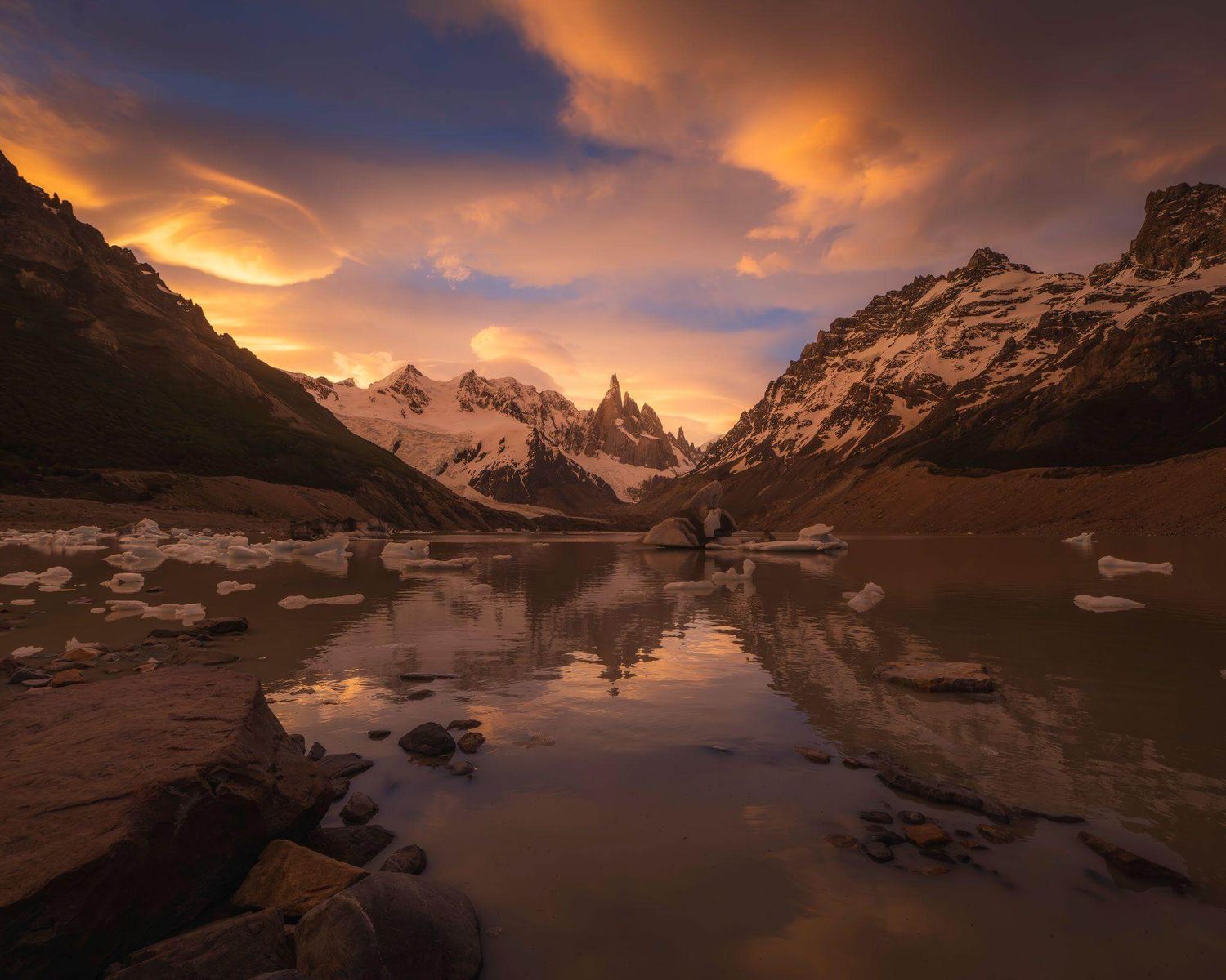 Fiery sunset clouds reflect in glacial lake beside snow-covered Cerro Torre peaks in Los Glaciares National Park.