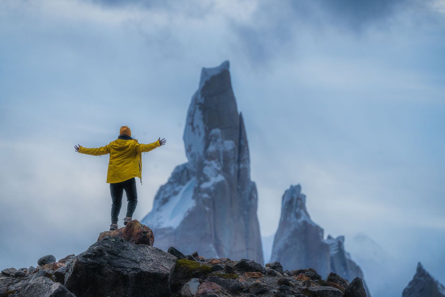 Person in yellow jacket standing on rocky peak with dramatic snow-capped mountain spires emerging through misty clouds.