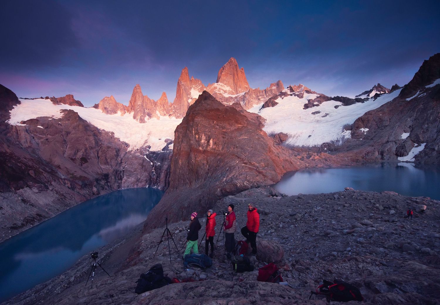 Hikers gather at twilight near a glacial lake with jagged snow-capped peaks of Mount Fitz Roy glowing at sunrise.