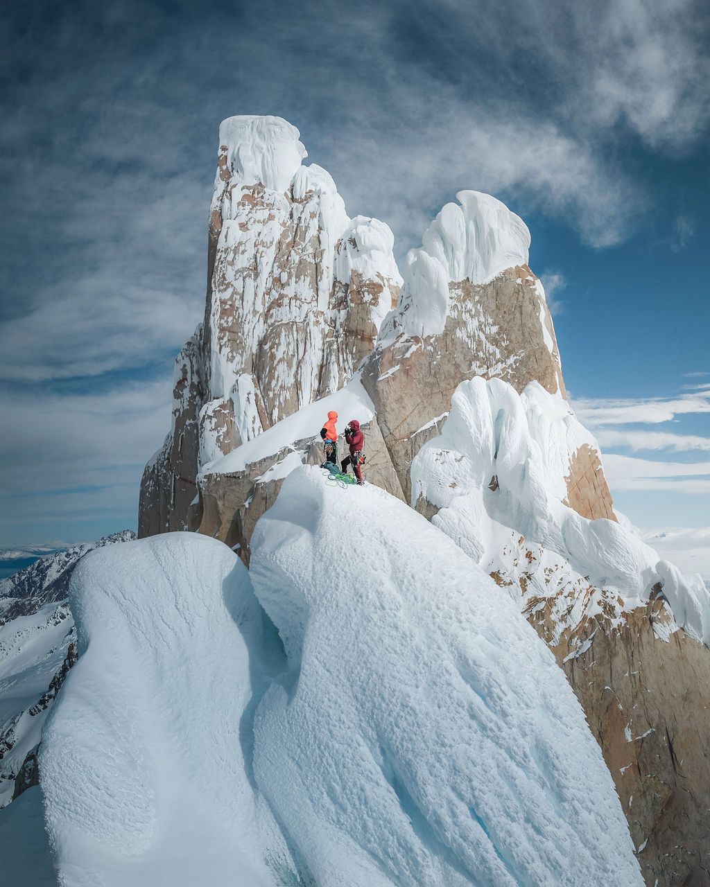 Two mountaineers standing among massive ice formations on a snow-covered rocky summit under blue skies.