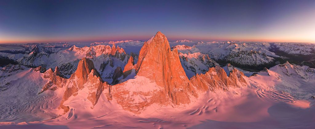 Wide aerial panorama of Fitz Roy massif glowing pink and orange at sunset over snow-covered Patagonian landscape.