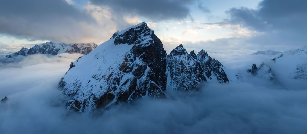 Snow-capped mountain peaks emerge dramatically above a sea of clouds at dusk in an aerial panoramic view.