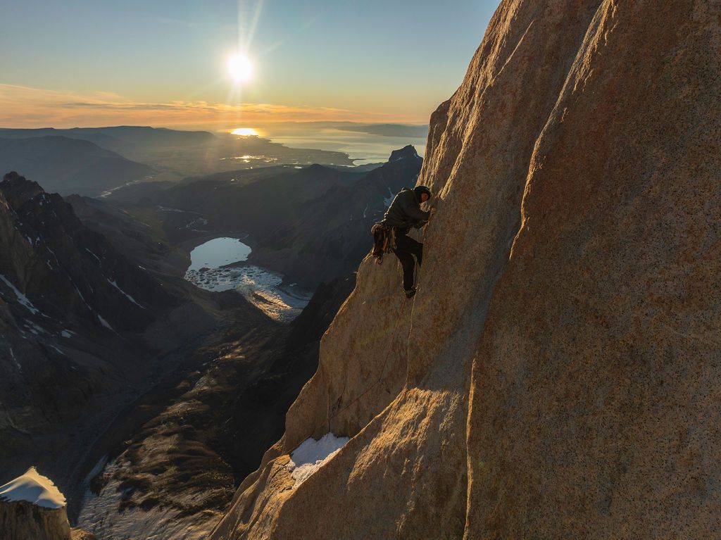 Climber on a sheer rock face at sunset with a glowing lake and vast mountain landscape below in Patagonia.