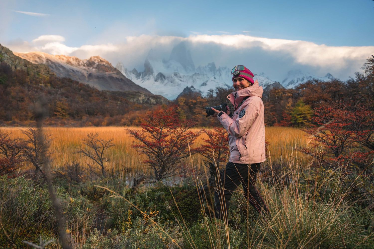 Woman in pink hat checks phone amid autumn foliage with snow-dusted Fitz Roy massif behind her in Patagonia.