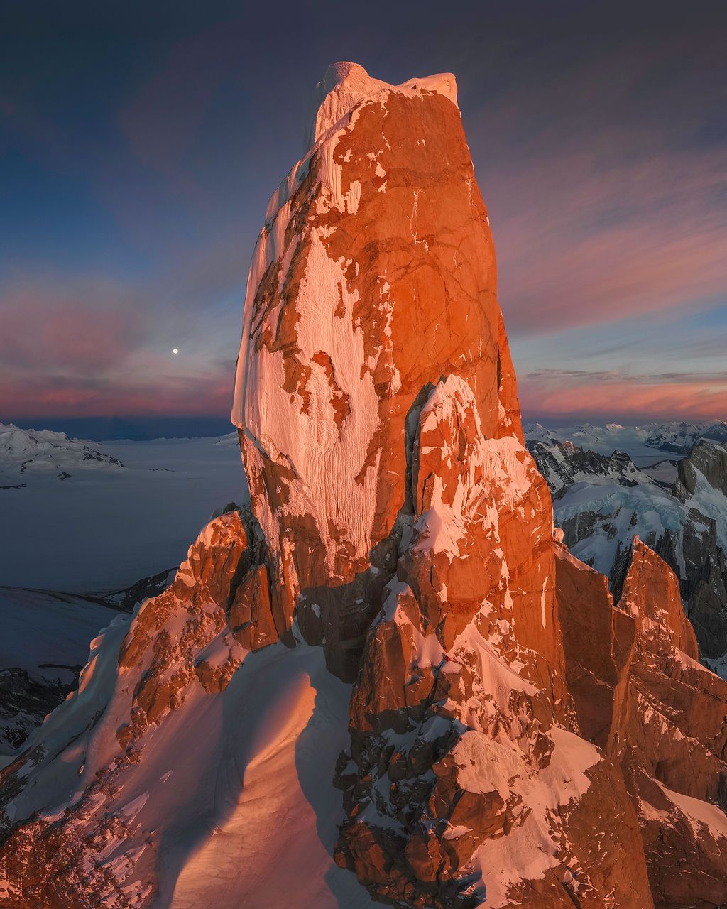 A massive red-hued granite tower glows in alpenglow light with a crescent moon above snowy peaks.