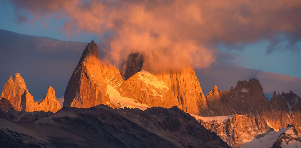 Mount Fitz Roy's iconic peaks glow vivid orange at sunrise beneath dramatic swirling clouds.