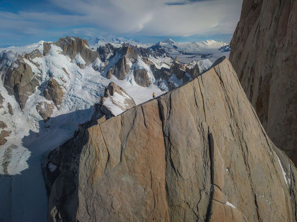 Aerial view of sheer granite rock face with snow-covered mountain range and glaciers in Patagonia below.