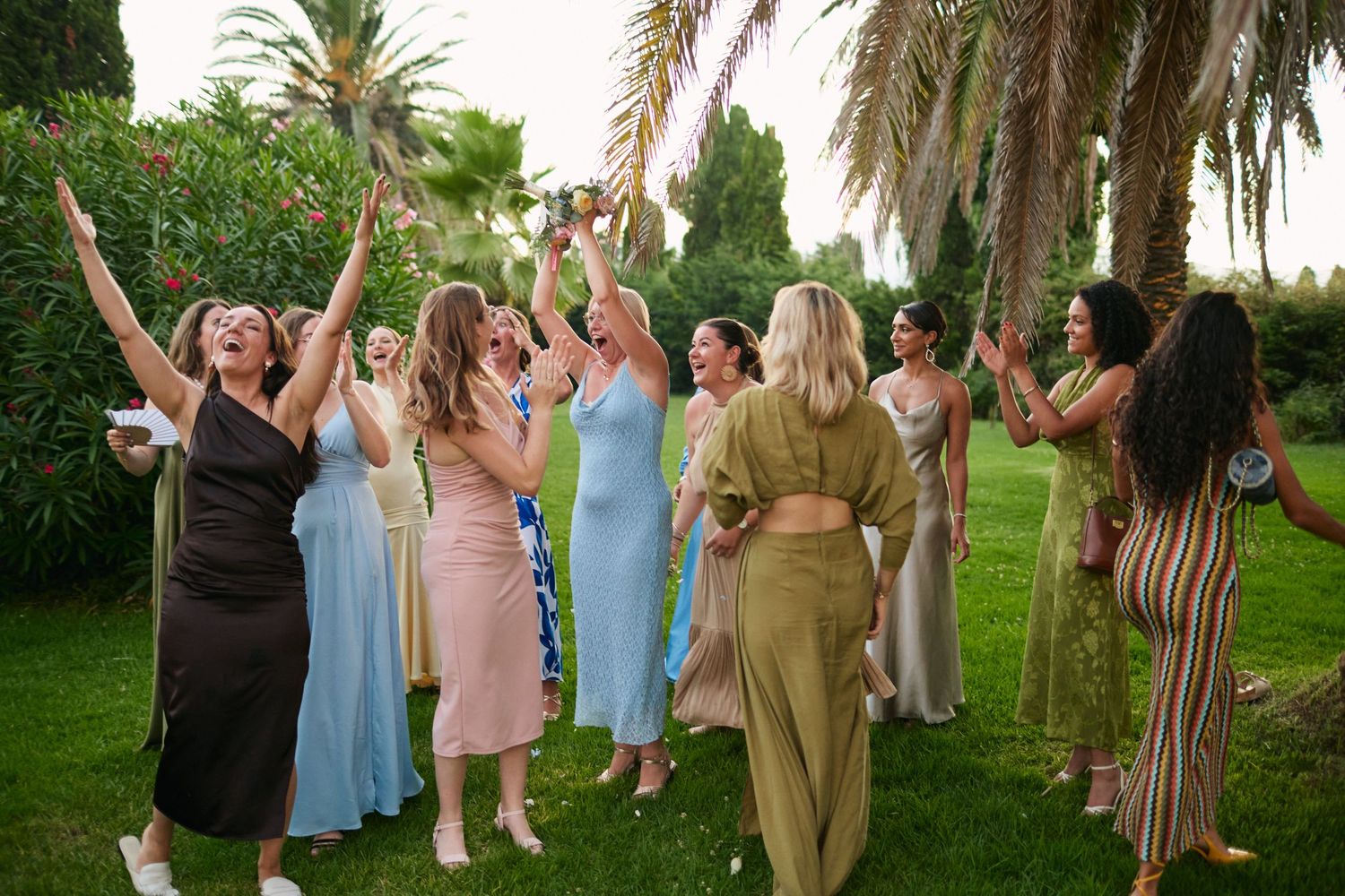 Wedding guests in elegant dresses cheering and celebrating bouquet toss outdoors near palm trees.