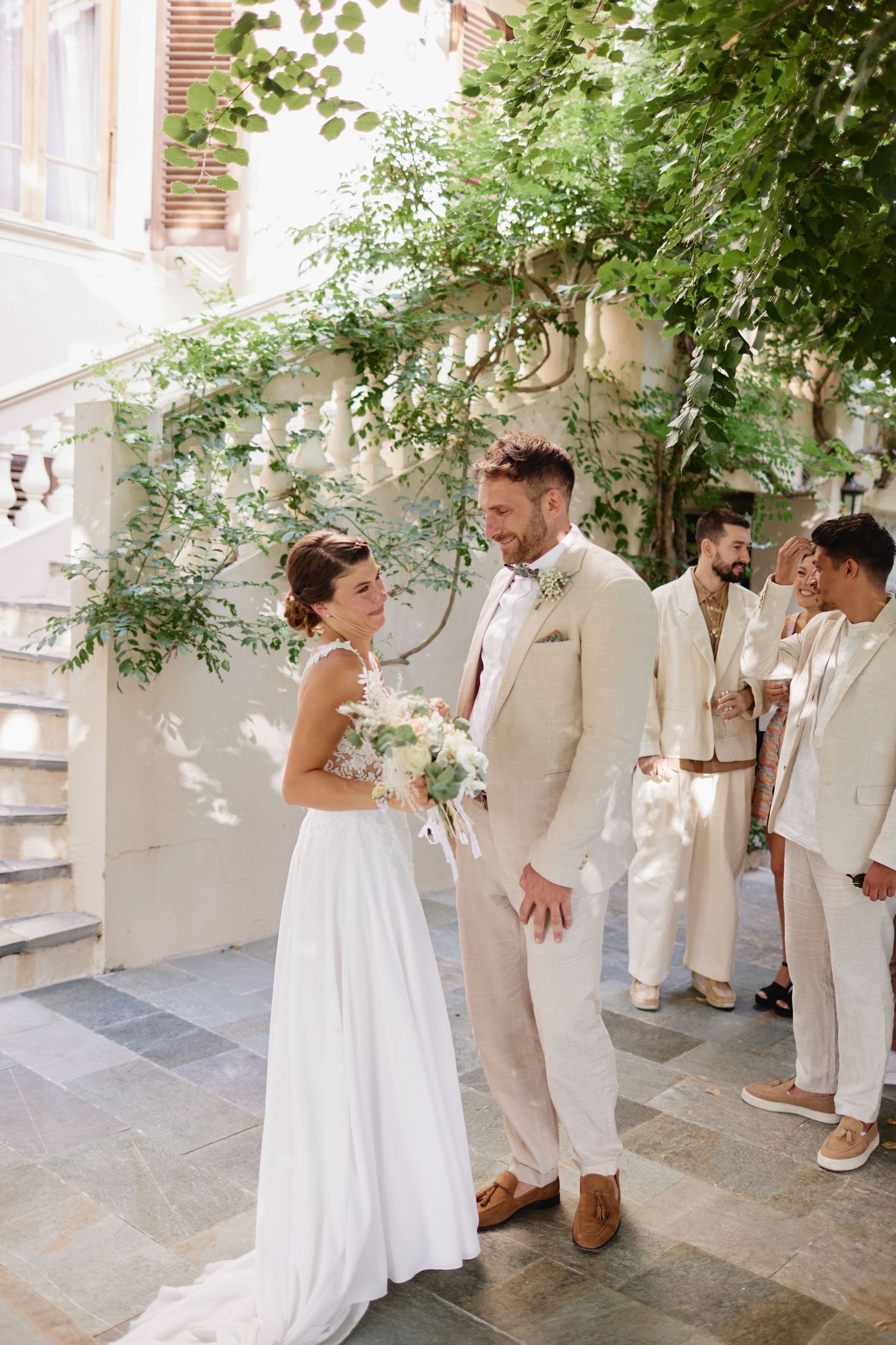 Bride and groom share a moment outdoors with groomsmen in beige suits in background.