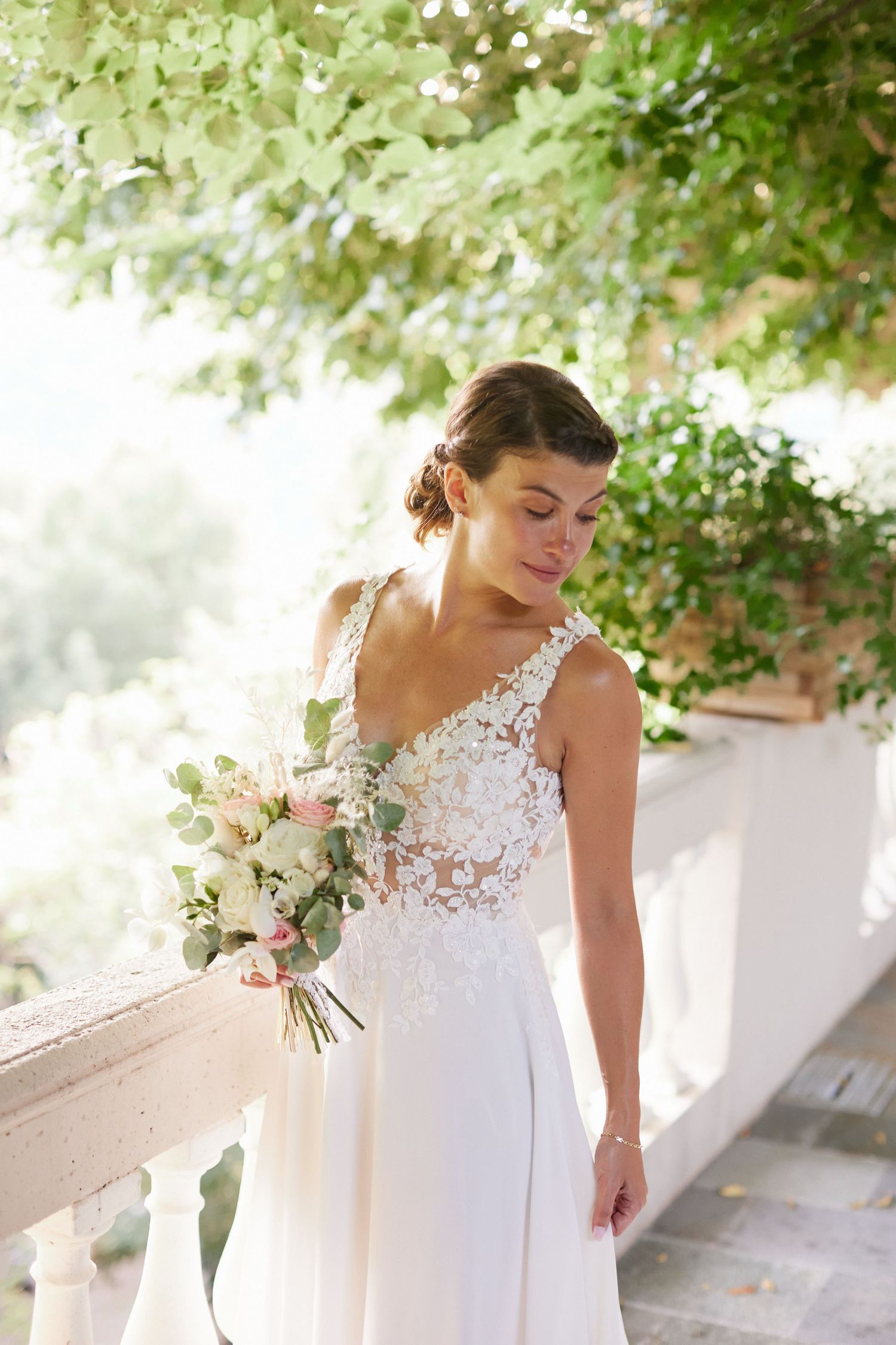 Beautiful bride in lace V-neck wedding gown holds pink and white bouquet outdoors.