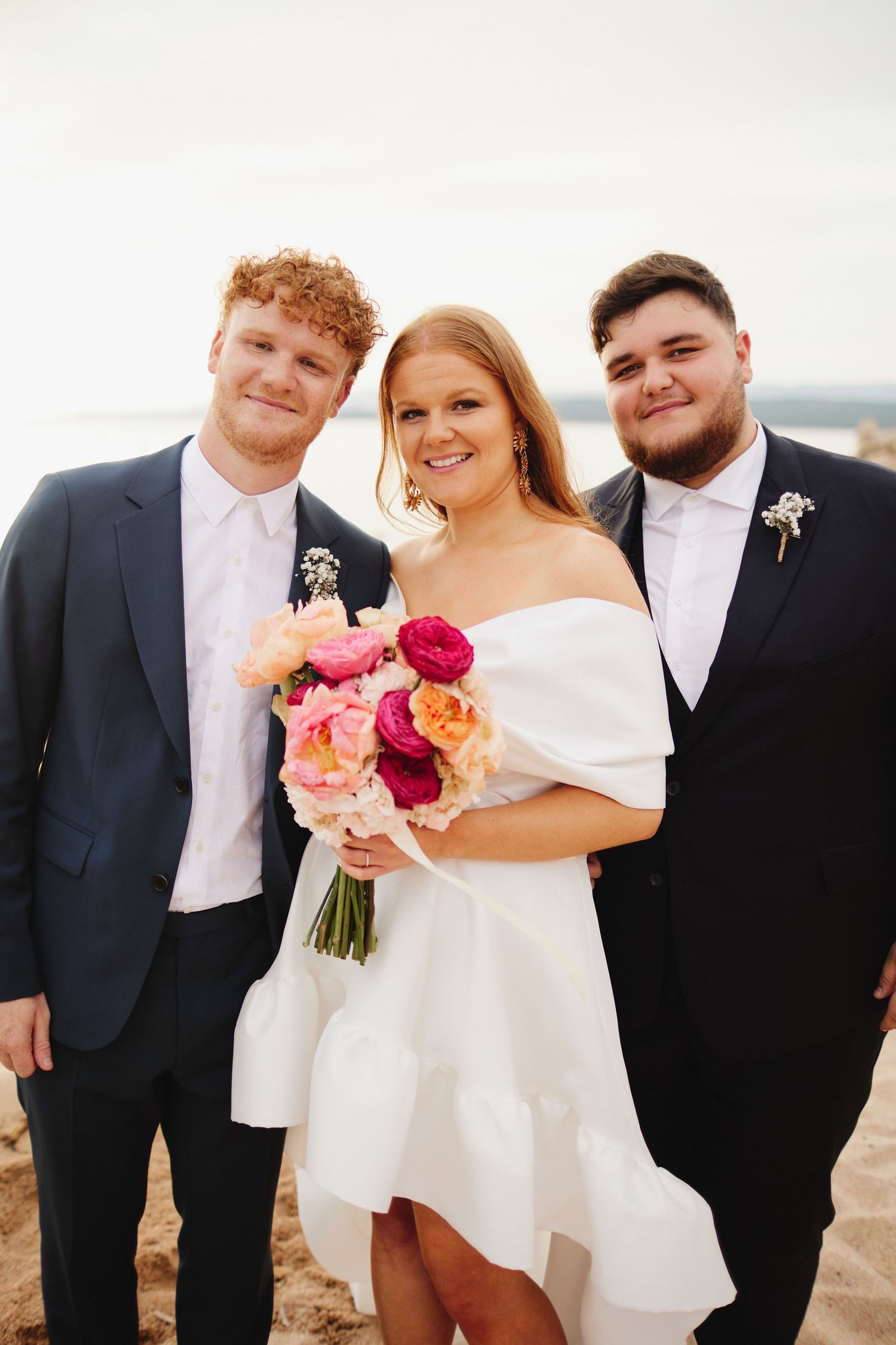 Smiling bride in off-shoulder white dress holding pink bouquet flanked by two groomsmen on beach.