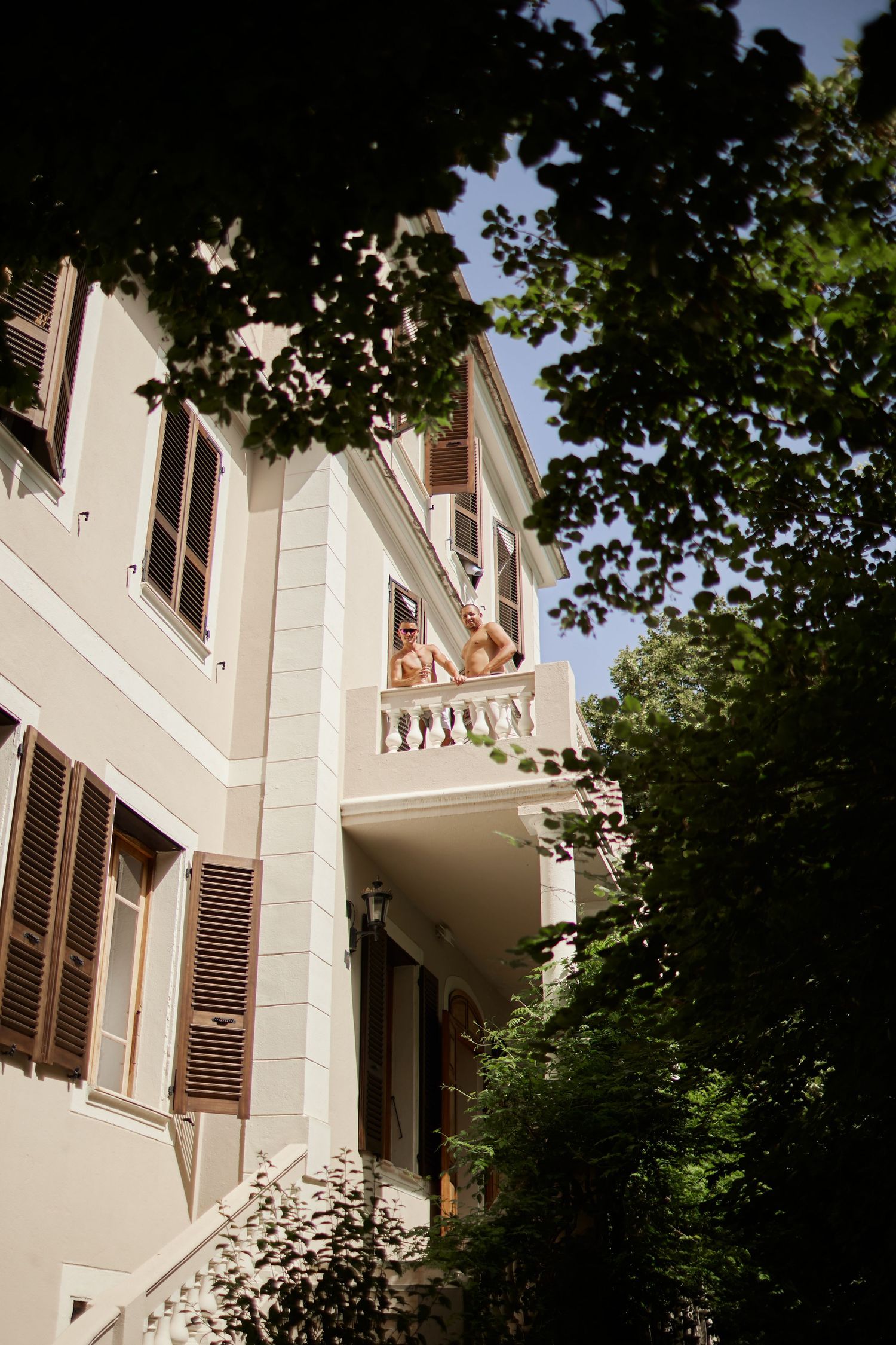 Two people standing on a balcony of a classic cream-colored villa surrounded by lush green trees.