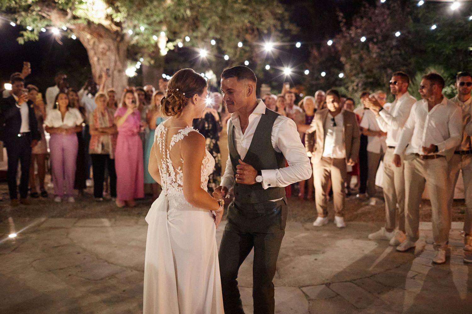 Bride and groom share first dance outdoors at night surrounded by wedding guests under string lights.