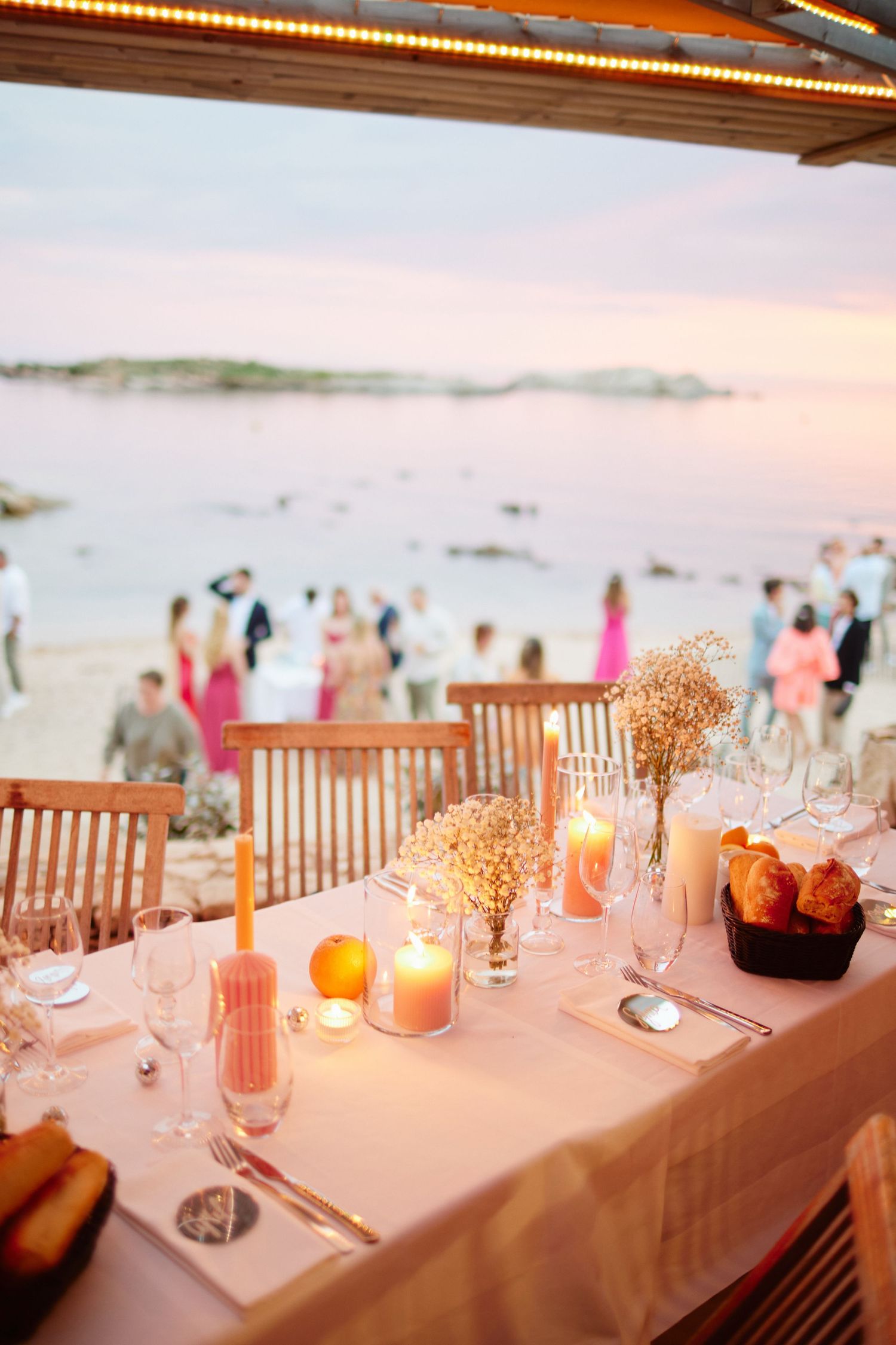 Romantic beach wedding reception table with candles, flowers, and guests mingling by the ocean at dusk.