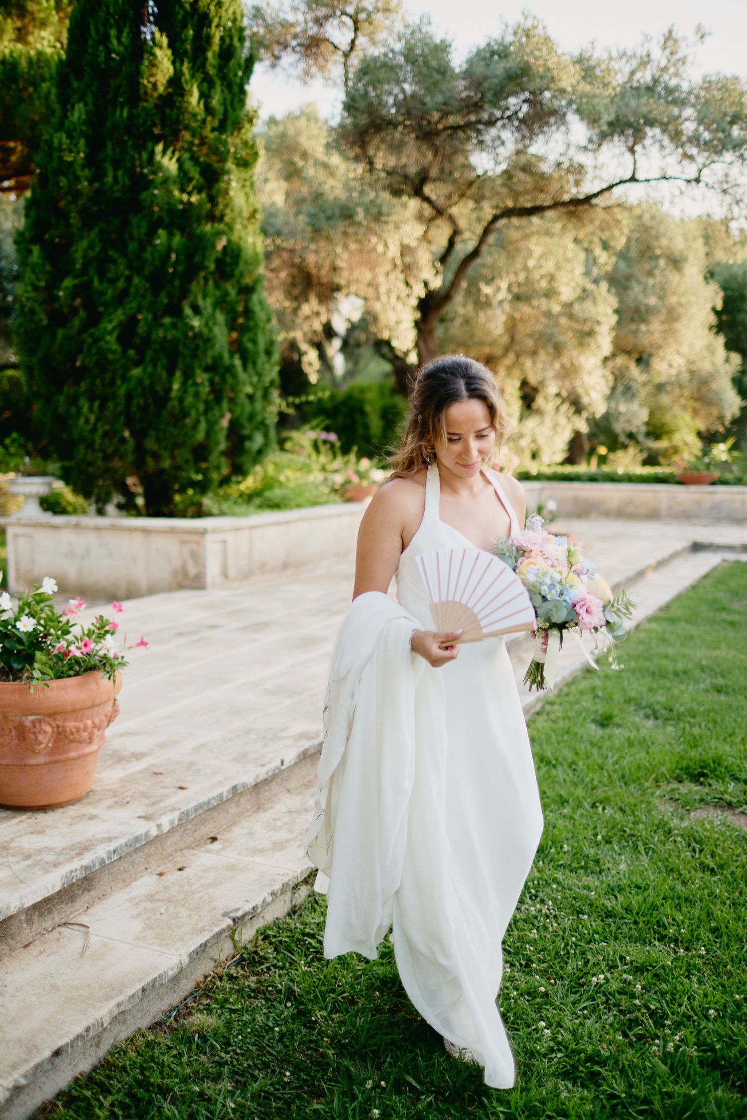 Bride in white halter gown holding bouquet and fan while walking through lush garden.