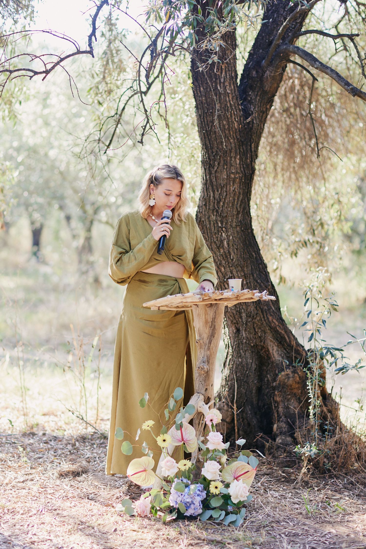 Female wedding officiant in olive green dress speaking at a rustic outdoor ceremony beneath a tree.