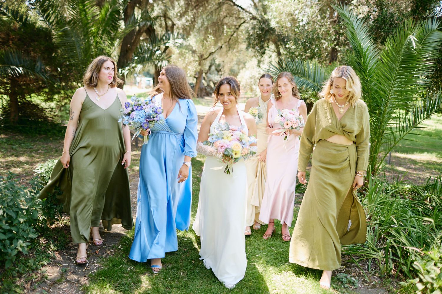 Bride and bridesmaids in pastel mismatched dresses walk together through a lush green outdoor garden.