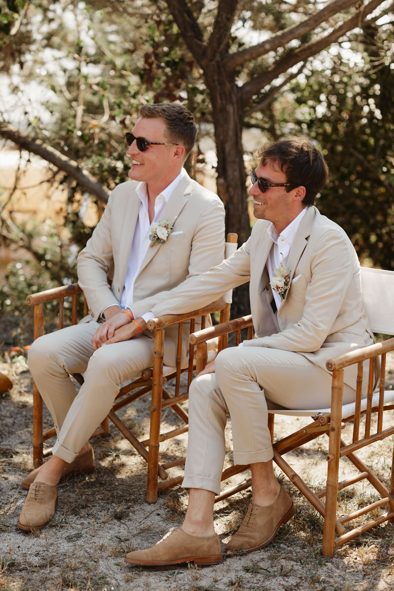 Two grooms in matching beige suits and sunglasses sit together at their outdoor wedding ceremony.