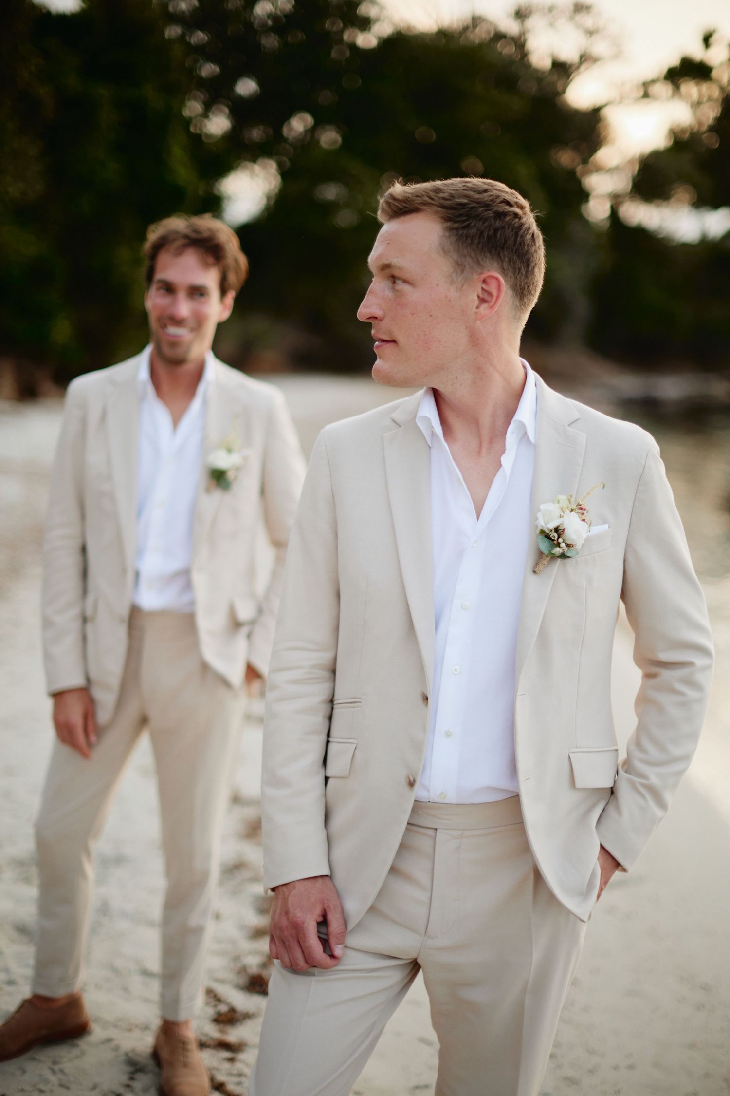 Two grooms in matching beige suits with boutonnieres pose on a sandy beach at sunset.