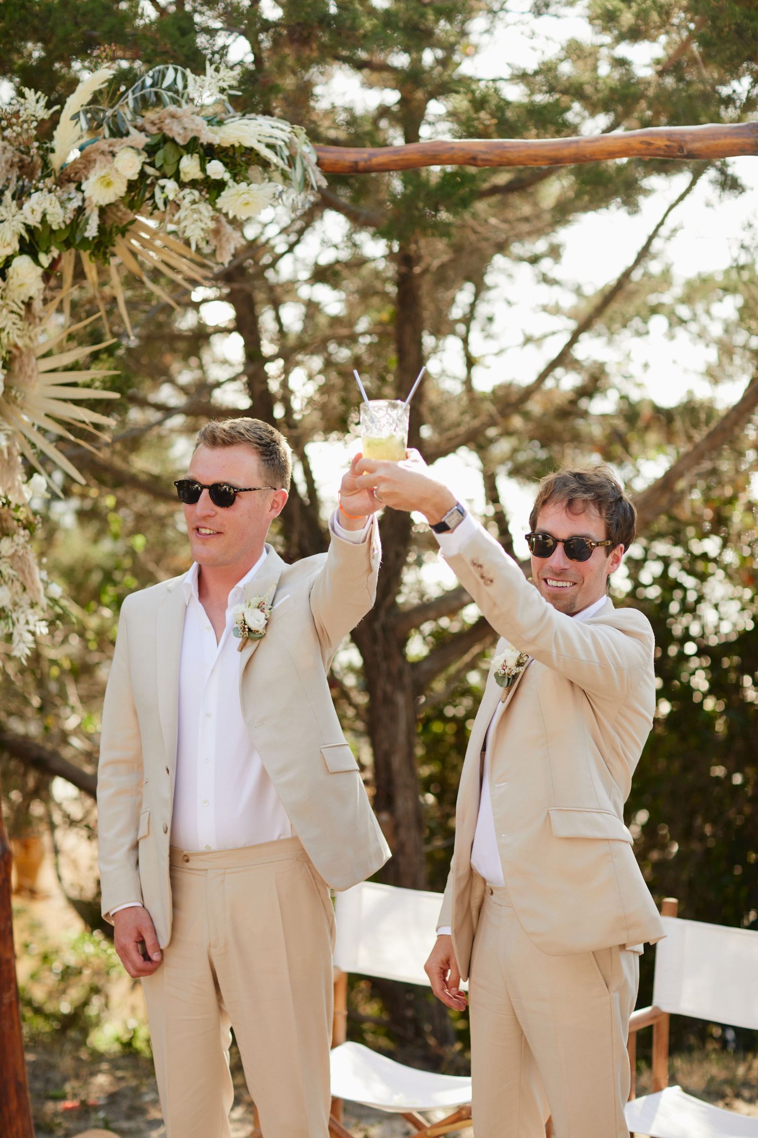 Two grooms in beige suits raise cocktail glasses in a toast at their outdoor wedding ceremony.
