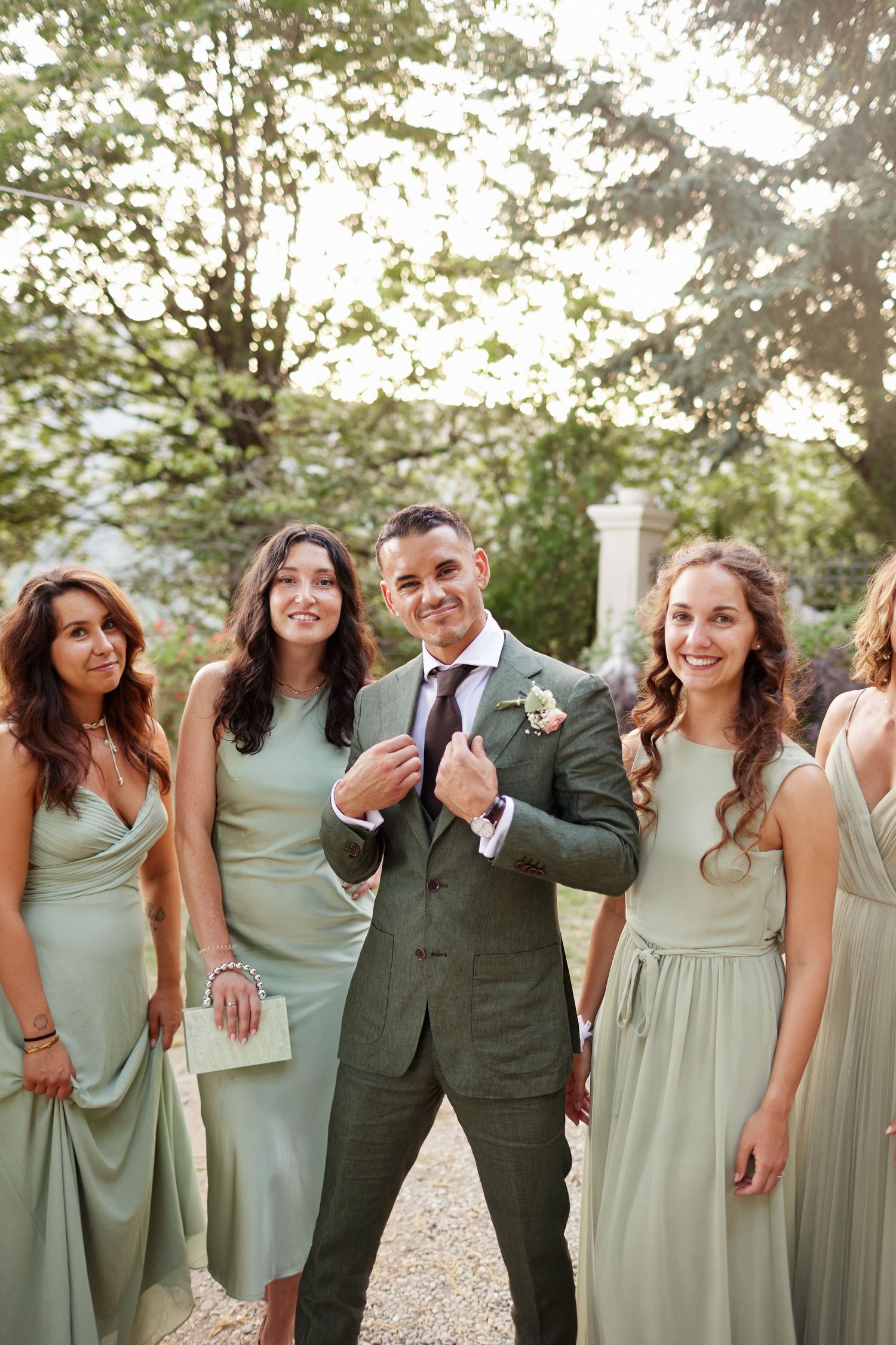 Groom in green suit poses confidently with smiling bridesmaids in sage green dresses.