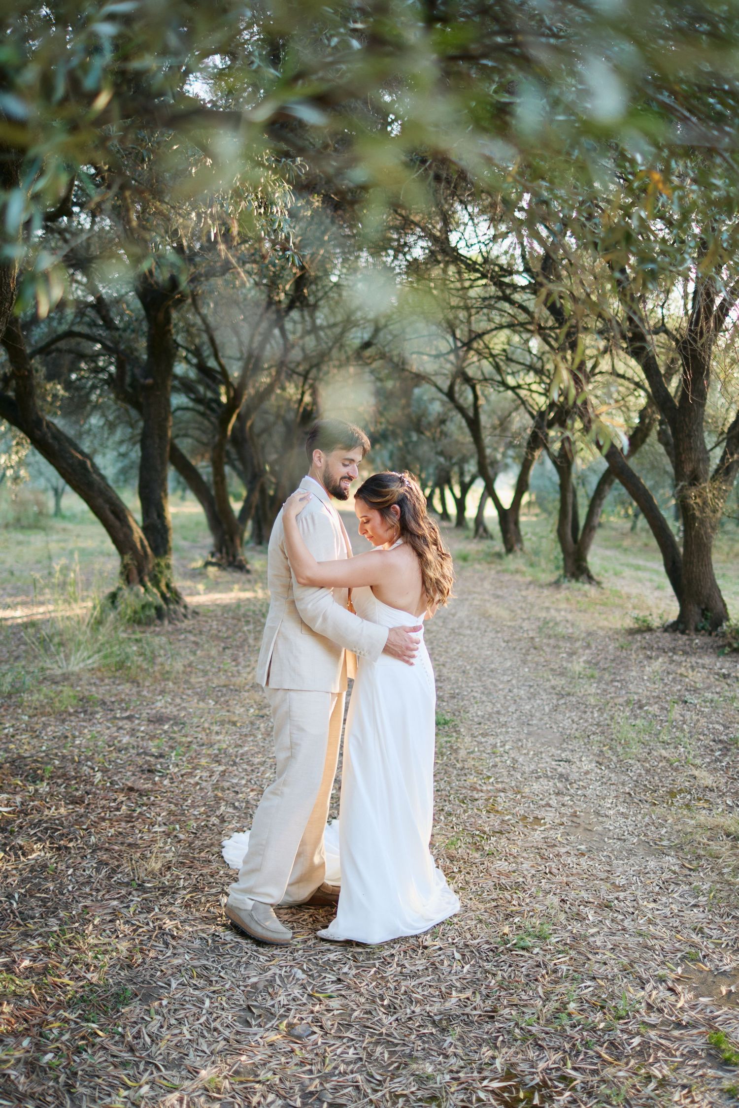 Groom embraces bride tenderly on rustic path through olive grove.