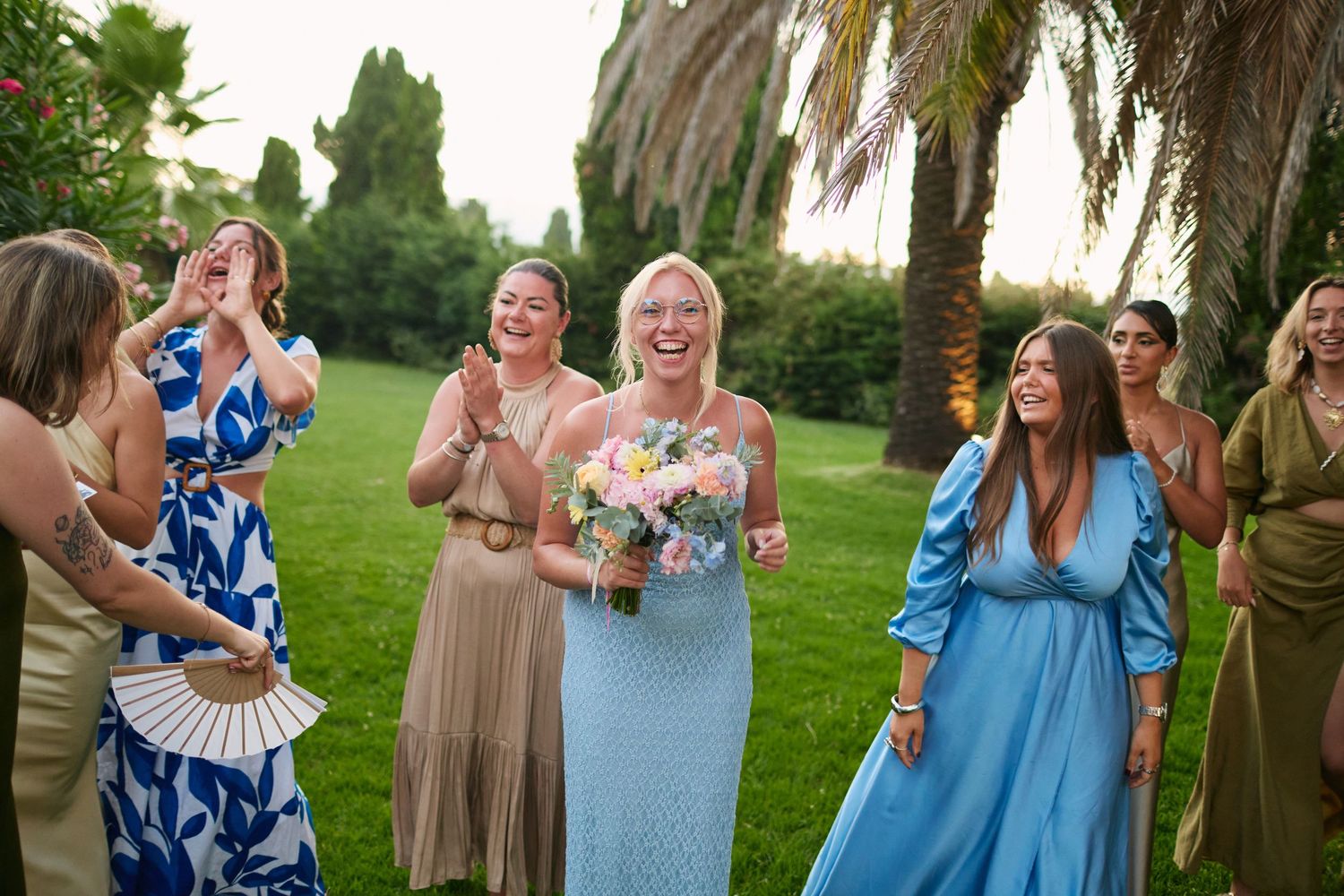 Happy wedding guest in blue dress holding bouquet surrounded by cheering friends at outdoor reception.