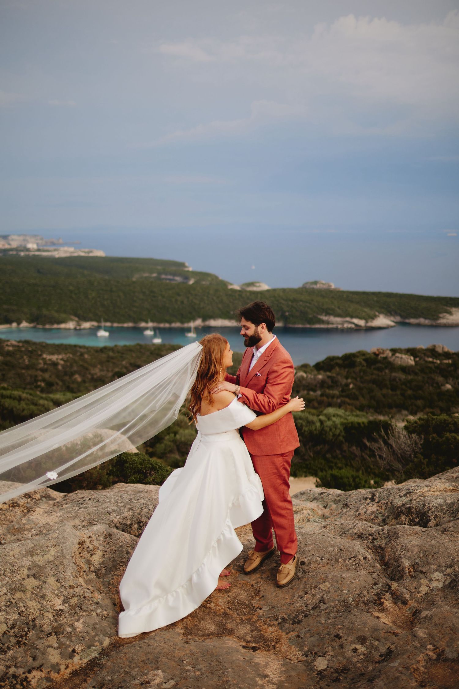 Bride's veil flows in the wind as couple embraces on rocky cliffs overlooking a turquoise bay.