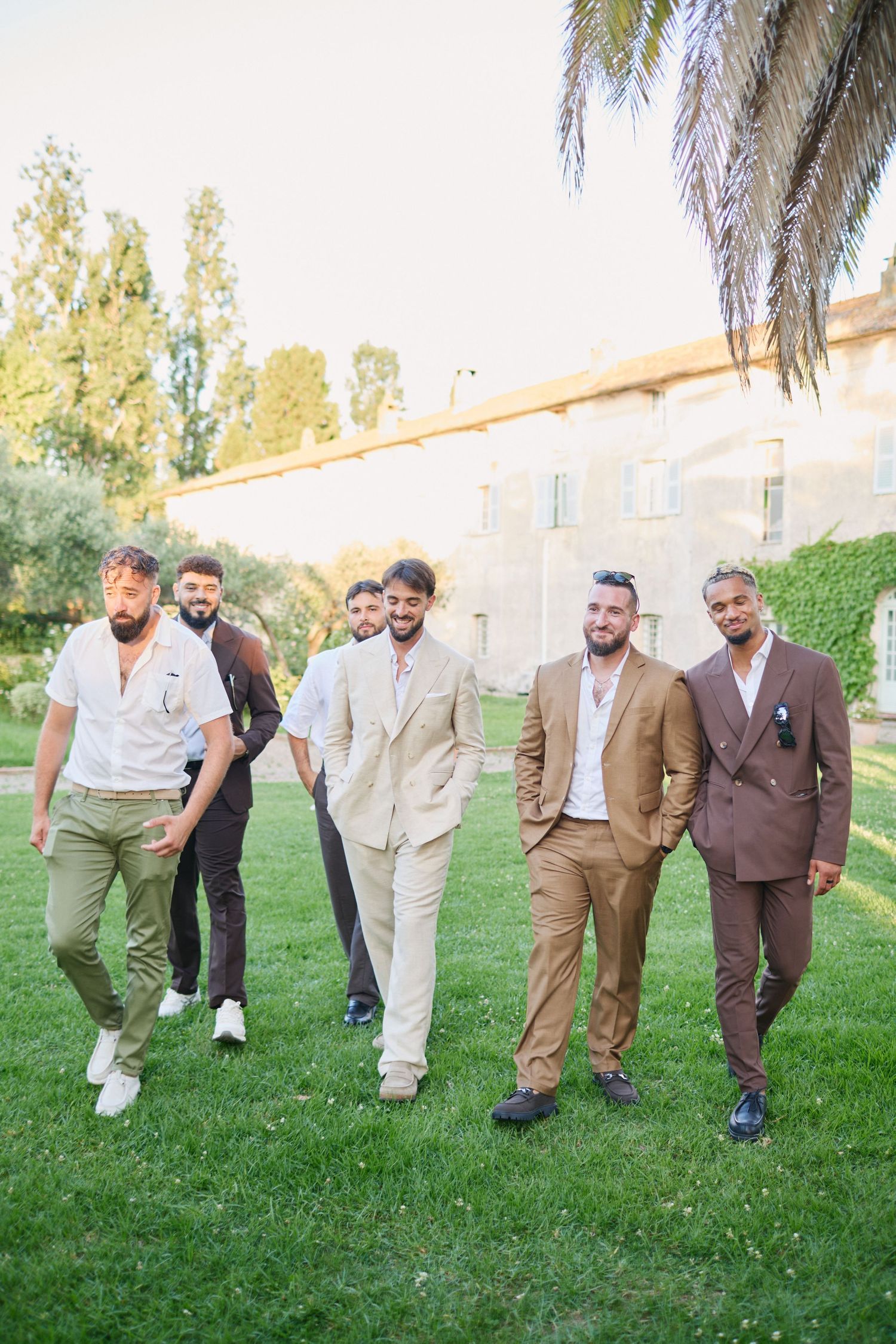 Groom leading groomsmen in suits walking confidently across lawn by chateau.