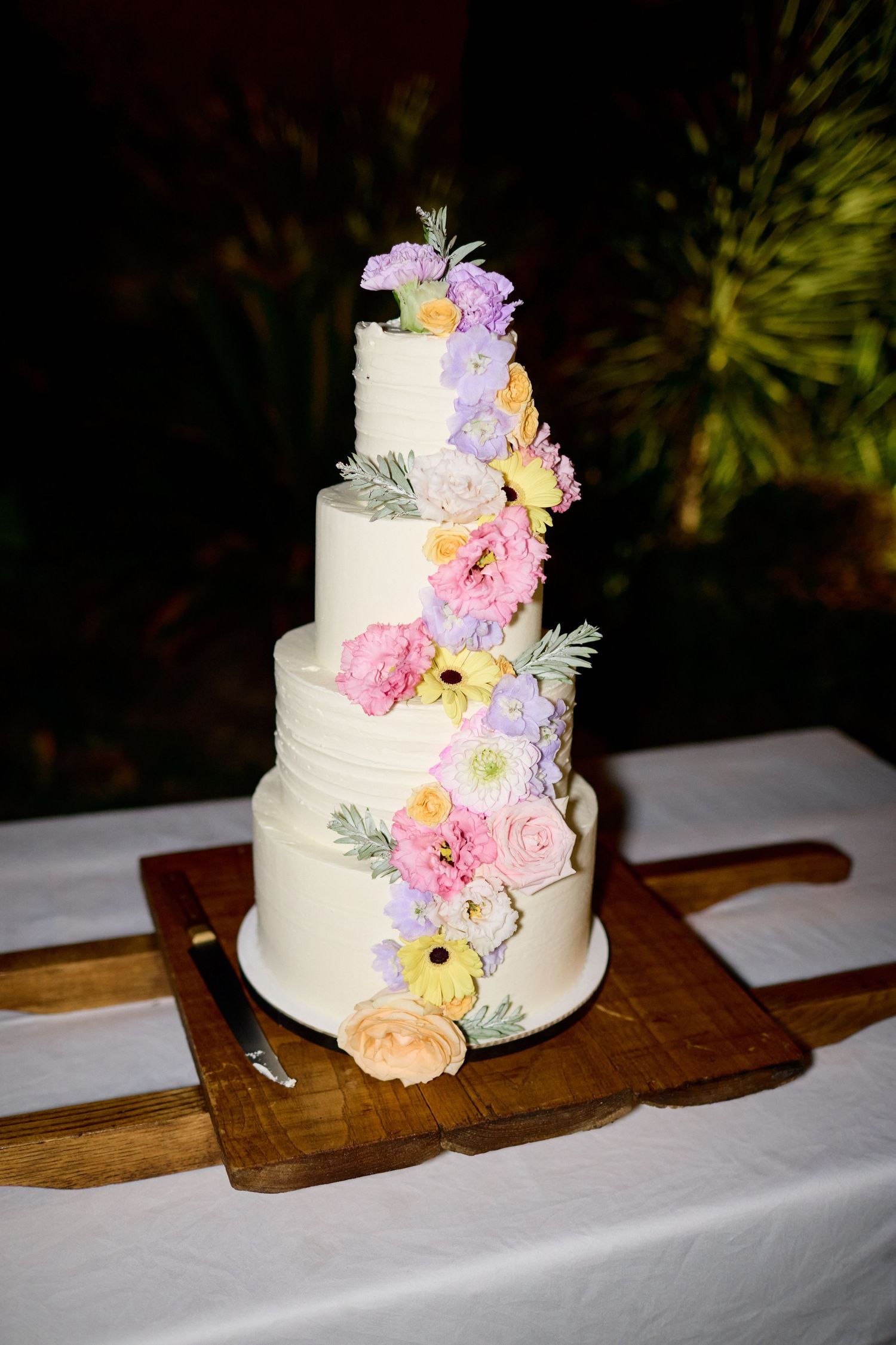 Elegant four-tier white wedding cake adorned with cascading colorful pastel flowers on wooden board.