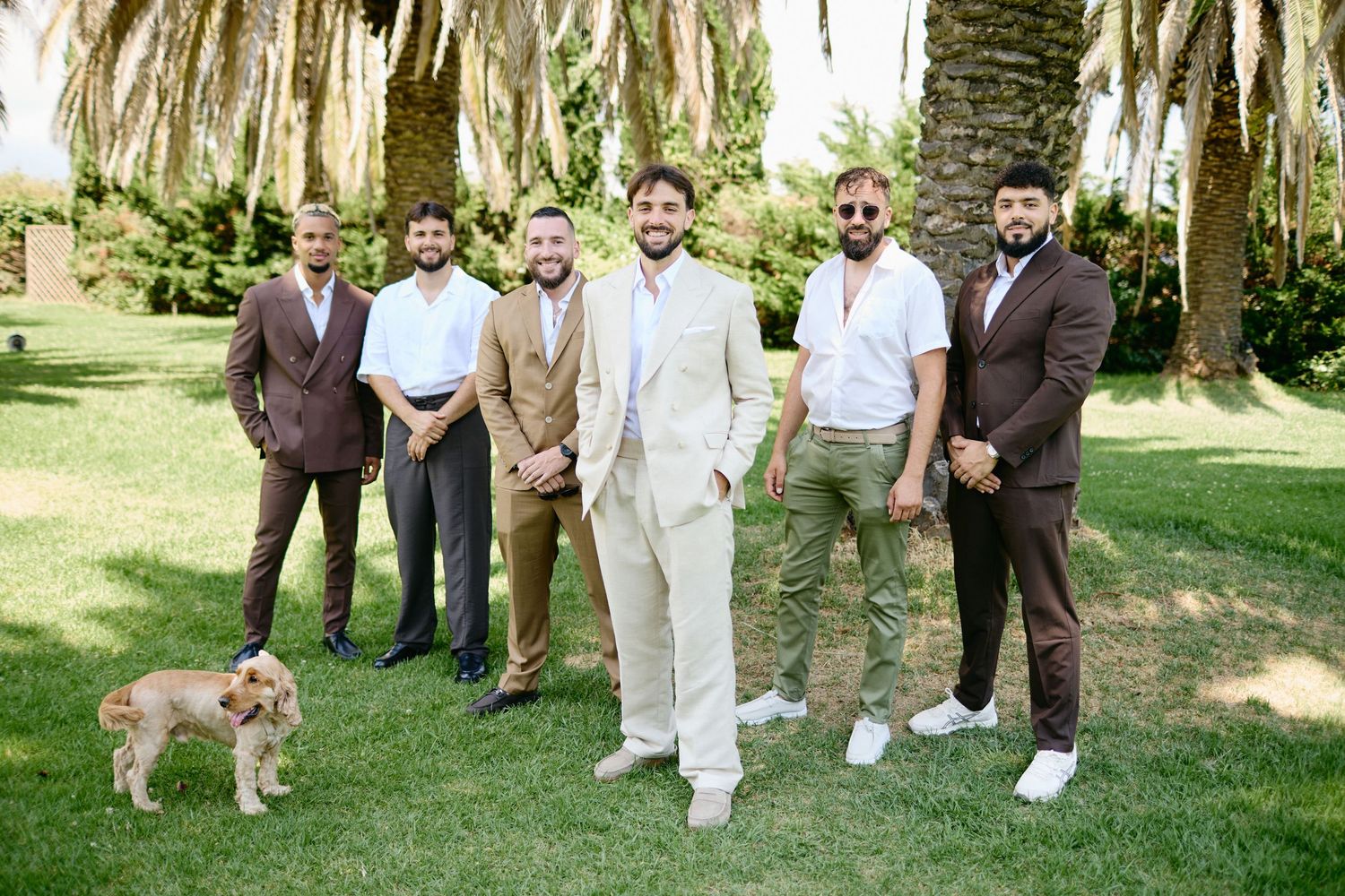Groom in beige suit poses with groomsmen in brown suits and a dog in a sunny garden.
