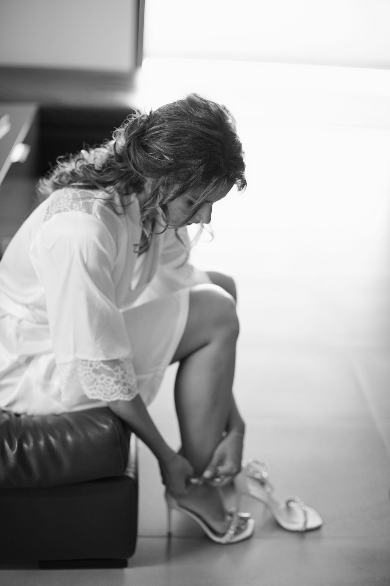 Black and white photo of bride in white robe putting on heeled sandals while seated before wedding ceremony.
